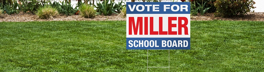 Red white and blue yard sign with text Vote Miller for School Board stuck in grass using wire stakes